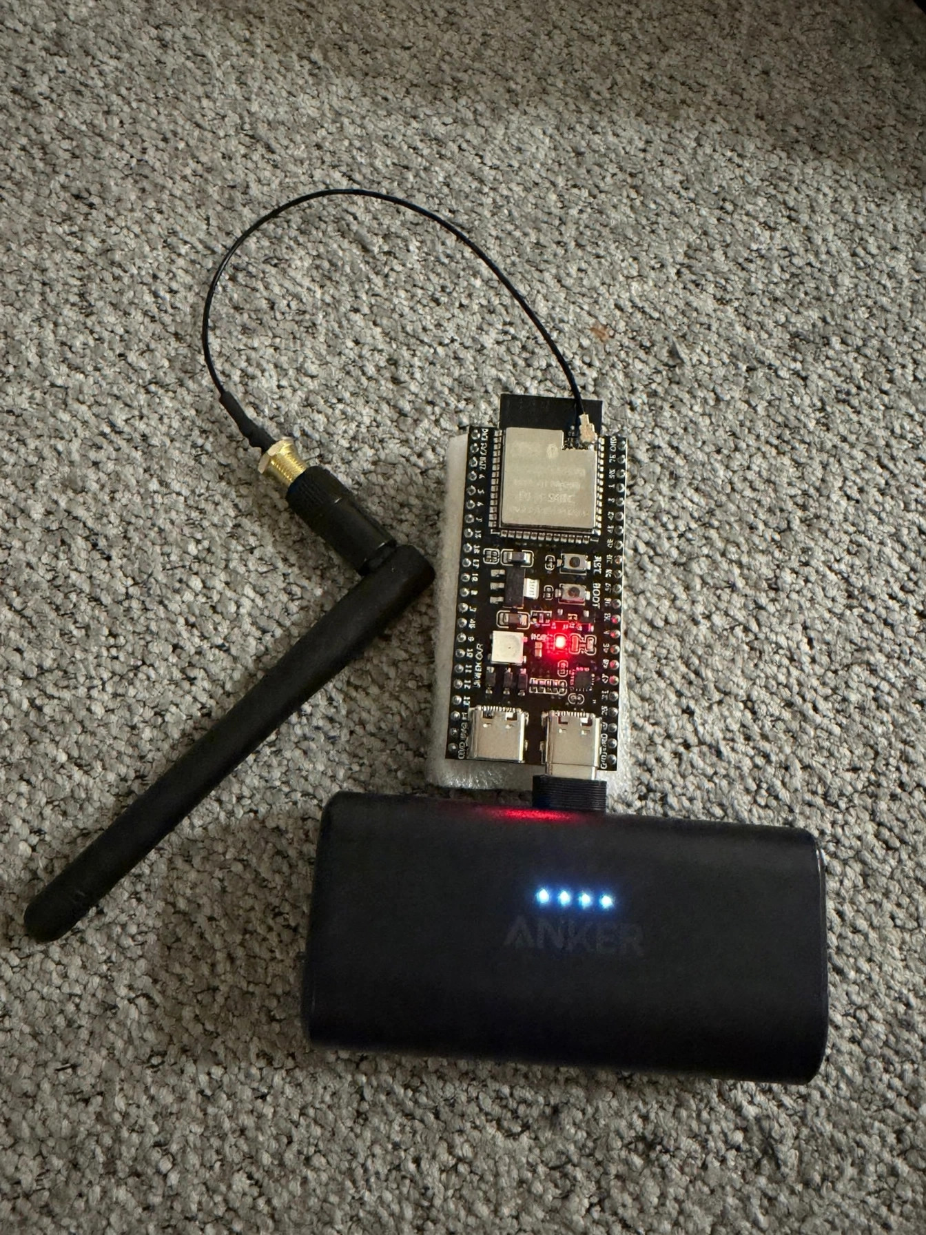 Herald's circuit board on the carpet, red LED lit, antenna connected, plugged into a power bank