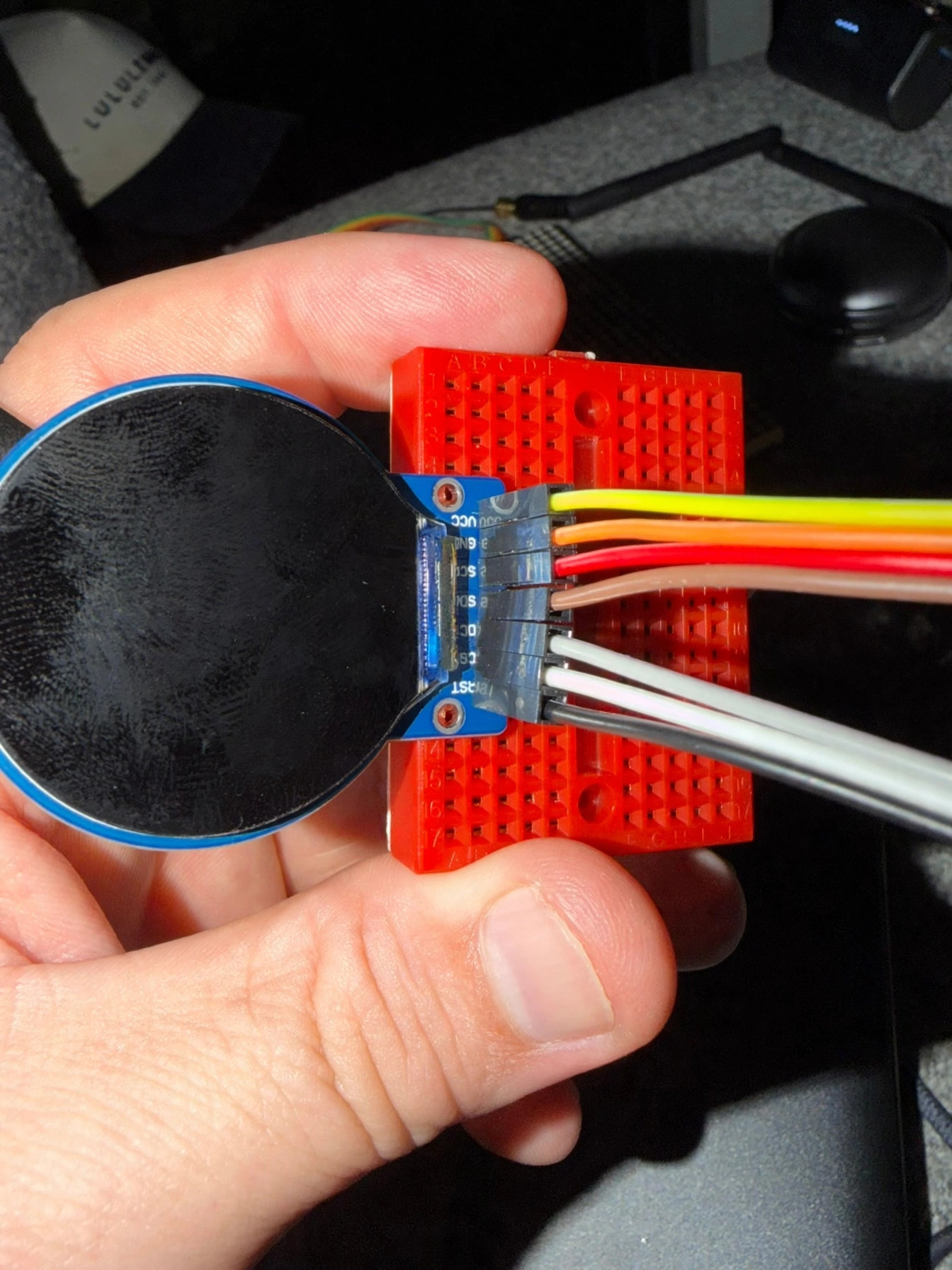 Locket's round display, dark, mounted on a red mini breadboard with colored and white wires, held in hand